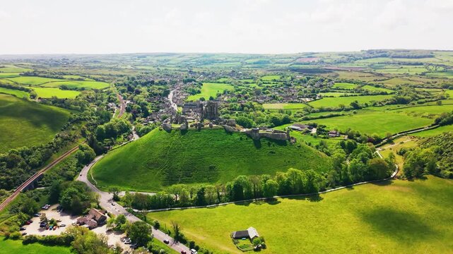 Aerial drone footage of Corfe Castle ruins standing on a green hill above the surrounding village and countryside in Dorset, England. The wide shot shows the historic castle, rural landscape, fields,
