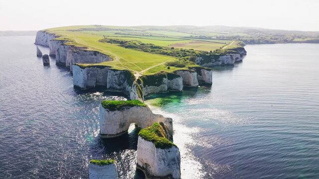 Wide aerial drone footage of Old Harry Rocks on the Dorset coastline in England, showing white chalk cliffs, sea stacks, a natural sea arch, turquoise water, green clifftop paths