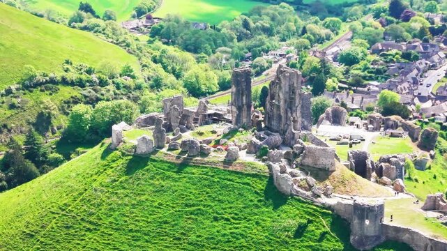Aerial drone footage of Corfe Castle ruins on a green hilltop in Dorset, England, with the surrounding village, roads, trees, and countryside visible below. The shot shows the historic medieval castle