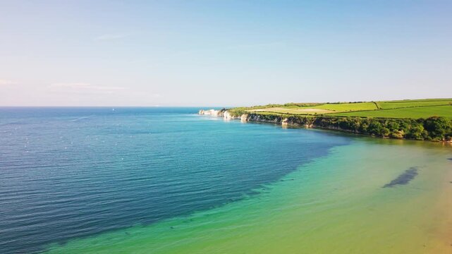 Aerial drone footage over clear turquoise sea along the Dorset coastline in England, with white chalk cliffs, green fields, calm water, and a bright blue sky.