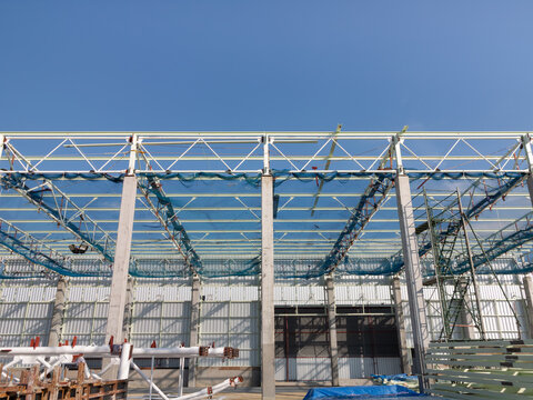 Industrial building under construction featuring concrete columns, metal roof truss frameworks and safety netting beneath  sky. Infrastructure development within factory or warehouse project.