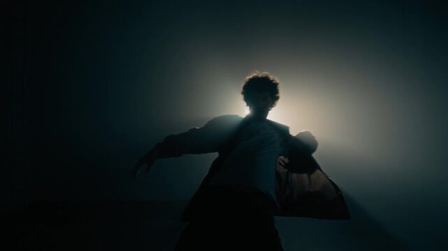 Artistic zoom out shot of young male dance performer silhouetted against glowing backlight highlighting fluid motion in dark studio, copy space