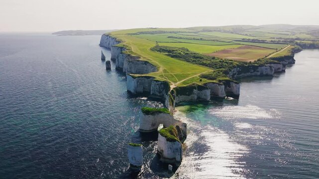 Aerial drone footage of Old Harry Rocks on the Dorset coast in England, showing a natural sea arch, white chalk cliffs, sea stacks, green clifftop paths, farmland, and calm blue sea.
