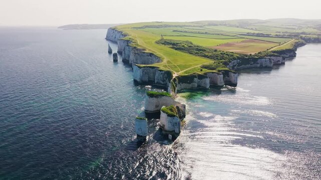 Wide aerial drone footage of Old Harry Rocks on the Dorset coast in England, showing dramatic white chalk sea stacks, coastal cliffs, a natural sea arch, green clifftop paths, farmland,