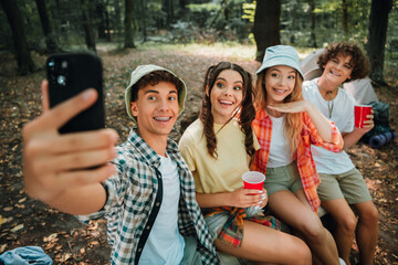 Four teenagers taking a happy group selfie while camping in the woods, smiling friends enjoying an...