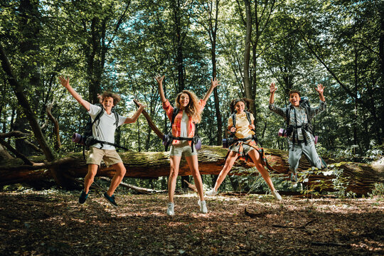 Four friends jumping with backpacks on a summer hiking adventure in a sunlit forest, laughing and celebrating group travel and outdoor fun