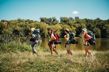 Naklejka premium Friends backpacking along a lakeside trail on a sunny summer day, hiking together with camping gear and sleeping mats in a scenic forest landscape