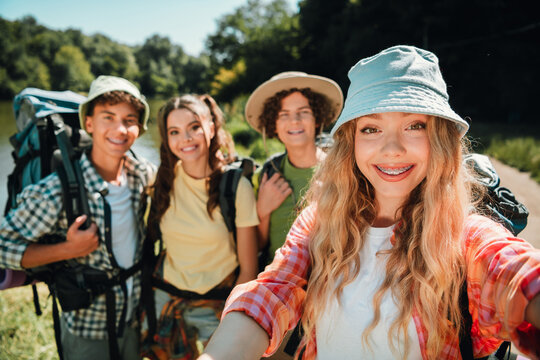 Smiling teenage friends taking a selfie on a sunny lakeside hike with backpacks and bucket hats, enjoying outdoor adventure, summer travel and carefree youth exploration