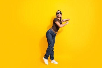 Fototapeta premium Smiling young woman in sunglasses posing on vibrant yellow background in casual jeans and sneakers expressing joy and playful energy