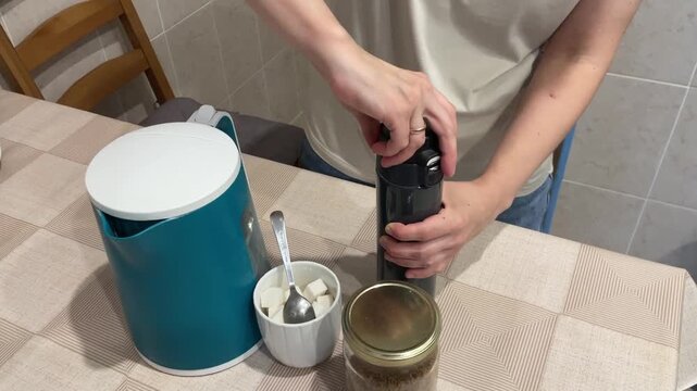 white woman filling thermos at table with kettle and sugar bowl, unscrewing lid and inspecting interior next to teal canister and ceramic cup, closeup hands on patterned tablecloth, warm domestic