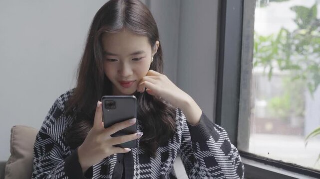 Young woman tapping smartphone by window, closeup expressions in cozy seat, soft natural light, patterned cardigan, scrolling