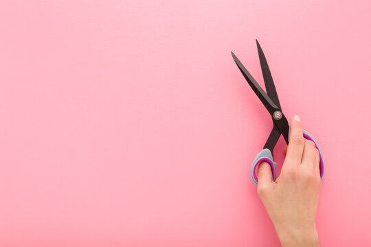 Young adult woman hand holding and showing new dark black dressmaker scissors for fabric cutting on light pink table background. Pastel color. Closeup. Empty place for text. Flat lay. Top down view.