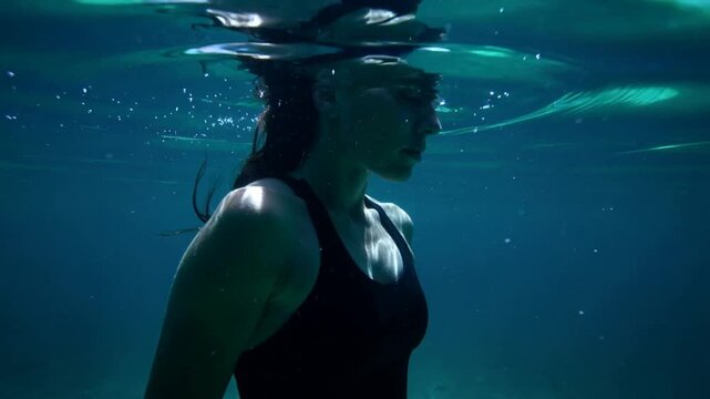 Floating fit female in black swimsuit holding vertical pose at water surface, with rising bubbles