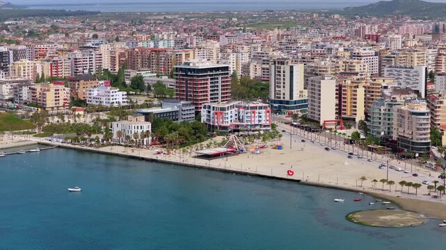 Aerial video of the waterfront promenade in Vlora, Albania, featuring clear turquoise sea water, green park pathways, and coastal hotel buildings.