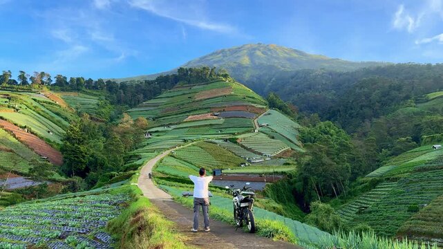 Man standing next to motorcycle overlooking terraced hills. Peaceful rural landscape with scenic nature view.