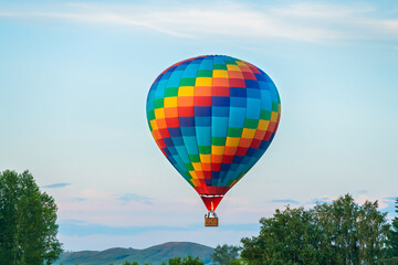 Obraz premium Colorful hot air balloon flying over the green hills and trees at sunset. Rainbow colored balloon in the blue sky.