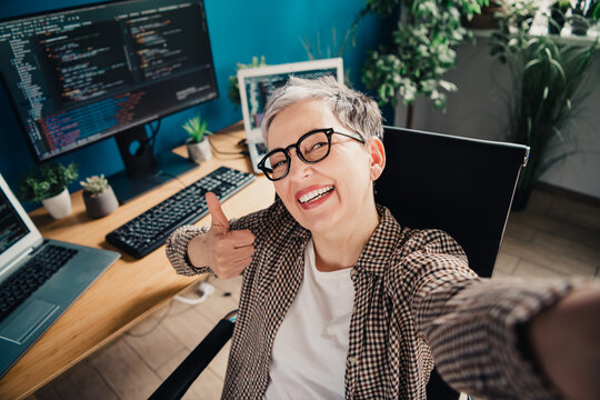 Smiling female programmer taking a selfie at her home office with monitors showing code, giving thumbs up while working remotely on software development
