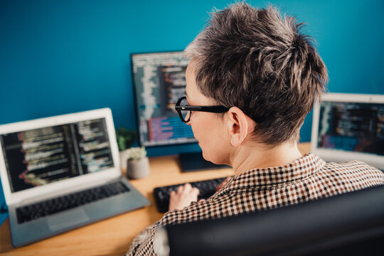 Mature female programmer coding at a multi-monitor home office desk, focused developer working on software with laptop and monitor in a modern remote work setup