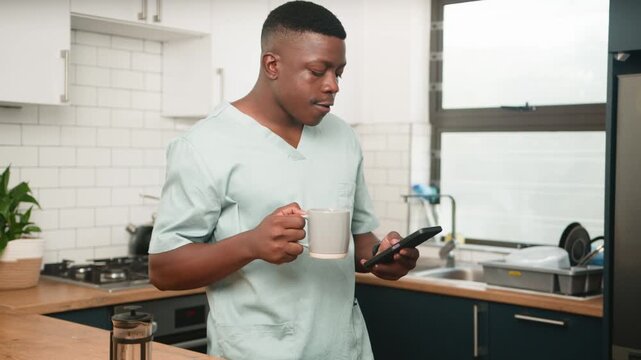 Young Black Man In Scrubs Drinks Coffee And Uses Phone In Modern Kitchen