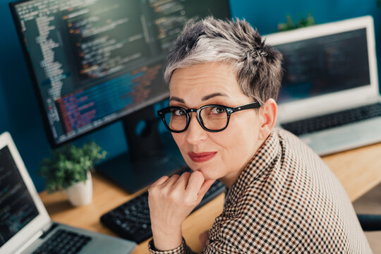Experienced female software developer with short gray hair and glasses working on code at a multi-monitor desk in a modern home office