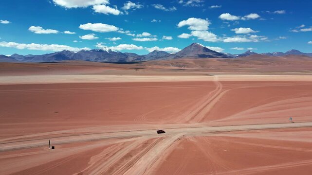 Dolly shot over pickup truck crossing a desert dirt road intersection on vast red altiplano plain with converging tire tracks and volcanic peaks ahead