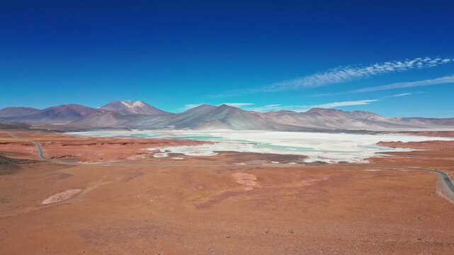 Panoramic aerial over vast red desert plain showcasing iconic turquoise lagoon and white salt flat framed by dramatic multicolored volcanic range. Piedras Rojas, Chile