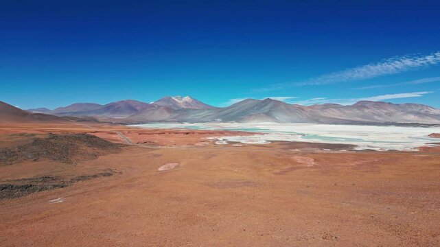 Ascending aerial over red arid altiplano desert revealing iconic turquoise lagoon and white salt flat surrounded by dramatic multicolored volcanic peaks. Piedras Rojas, Chile