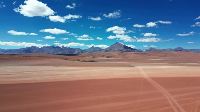 Slow backward flight over vast red arid altiplano desert plain with faint tire tracks revealing dramatic chain of volcanic peaks under vivid blue sky and clouds