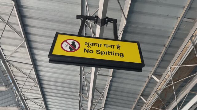 Dynamic orbiting shot of a yellow no spitting sign board hanging inside an Indian railway station with metal roof structure and public transportation environment.