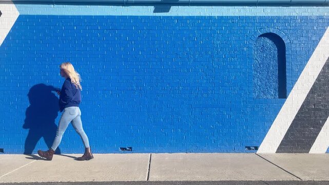 A woman is walking against a blue brick wall checking her phone app and stopping then walking again for a news update, announcement or email confirmation,