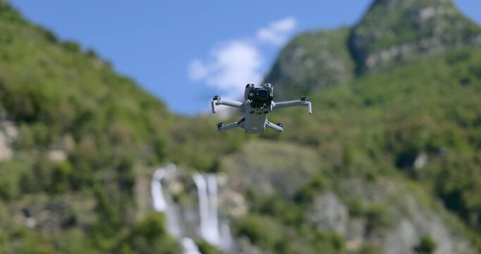 Close-up of DJI Mini series drone hovering in air with mountain waterfall and green forest in background. Rack focus