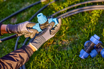 Fototapeta premium Closeup of hands in the gloves. Process of plumbing, installing fittings pipes and connectors for the garden and lawn irrigation system installation. Green grass, shallow depth of field.