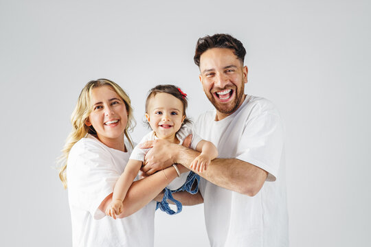 A cheerful family with a happy, baby. The parents are smiling and looking lovingly at their daughter. Everyone is wearing casual white tops and jeans.