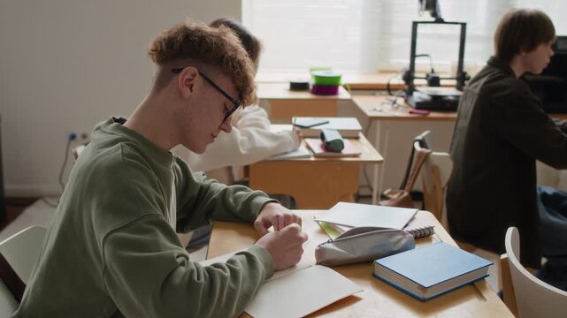 Curly haired teenage boy with large glasses taking notes in notebook while sitting at single-seat desk among other students in modern class