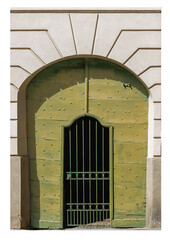 Obraz premium Antique green wooden arched door with embossed studded nails, with integrated pedestrian door protected by a wrought-iron grille