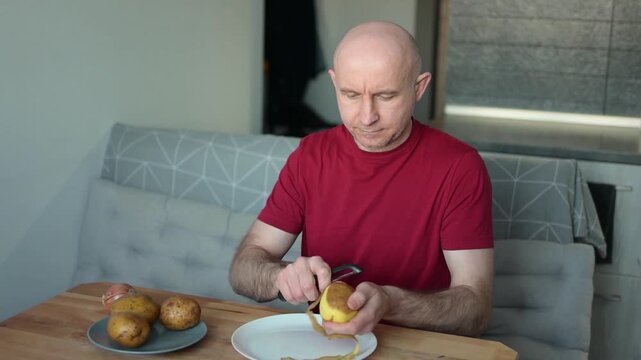 Bald man peeling raw potato with vegetable peeler in modern kitchen at home video Full HD