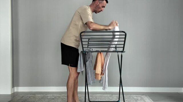 Barefoot man in casual clothing doing laundry and hanging clean wet clothes on a collapsible drying rack after washing. Domestic routine concept