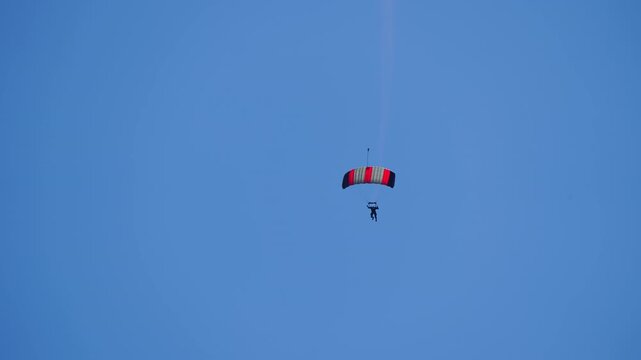 Aerobatic skydiver under a red-striped ram-air canopy performing a spiraling turn in clear blue sky, long white smoke trail marking the path