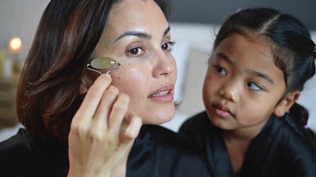 Bringing jade roller under eye, mom in black-satin robe showing skincare on bed, daughter watching