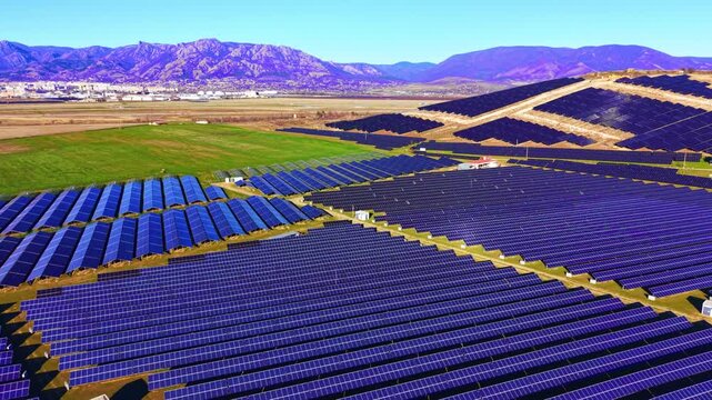 Tilted blue photovoltaic rows with green grass strips and gravel paths, small substation at center. High-angle view of solar panel array rows across open field, Bulgaria