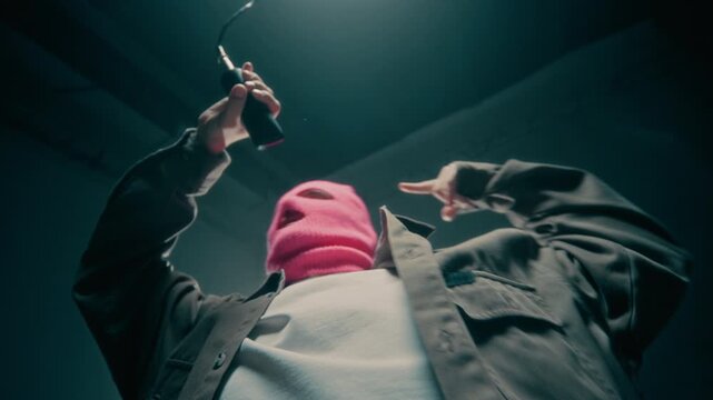 Dramatic low angle shot of masked rapper performing in music video singing to microphone in dark studio and gesturing aggressively, copy space