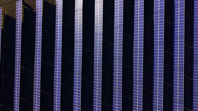 Close up pattern of vertical solar panel rows in sunlight. Aerial view of tightly spaced photovoltaic panels oriented vertically, alternating lit and dark striped texture in blue tones.