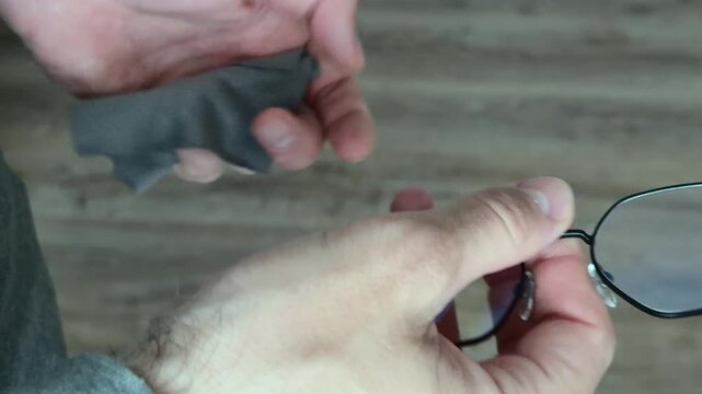 Close up of unrecognizable male hands meticulously cleaning eyeglasses with a microfibre cloth opening the frames checking lenses for spots against soft indoor daylight before putting them on.