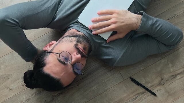 Contemplative writer lying on floor with notebook on chest, tapping page while thinking through ideas, glasses on and pen nearby in window light, relaxed creative process from high angle at home