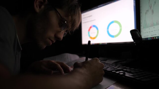 Male trader analyzing stock graphs on multiple computer screens and making notes on paper. Young financial analyst working with commodities and foreign exchange charts at night. Bitcoin BTC concept