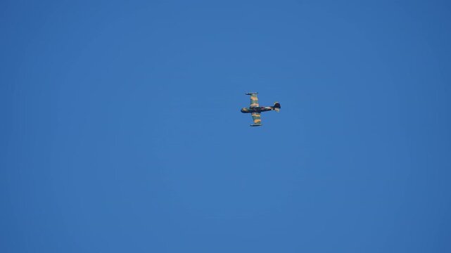 Military propeller trainer aircraft in camouflage livery performing a banking maneuver seen from below against a clear deep blue sky