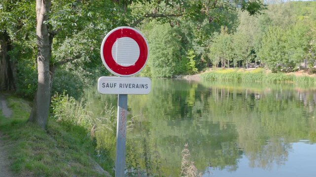 French road sign forbidding entry except for residents, located next to a peaceful river lined with lush green trees