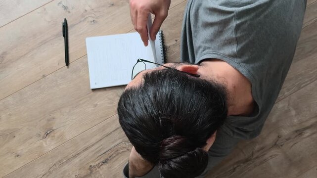 Unrecognizable long haired man in glasses reading handwritten notes in notebook while lying on side on wooden floor, calm slow life reflection, personal planning and creative review from above