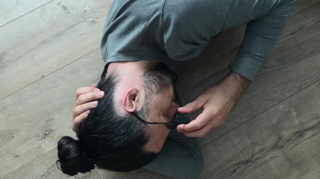 Withdrawn man lying on his side on wooden laminate floor removes glasses, places them beside him and rests hand under head, closed lonely pose showing emotional burnout in quiet overhead home portrait