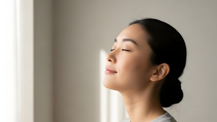 Young Asian woman relaxing, breathing deeply with eyes closed, enjoying sunlight near window